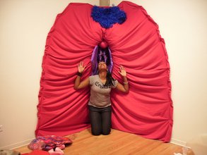 A woman in a worship pose on her knees, hands halfway raised and eyes are closed. Behind her is a bright pink cloth draped into a labia with curly royal blue pubic hair. The woman's head sits right in the center of a vulva made of purple fabric.