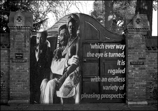 A black and white photo of a large wooden gate flanked by brick pillars and wall. An image of a man smoking on an armchair sits next to a sailor with a shirtless woman wearing a headscarf on his lap. Large white text is typed on the right gate door.