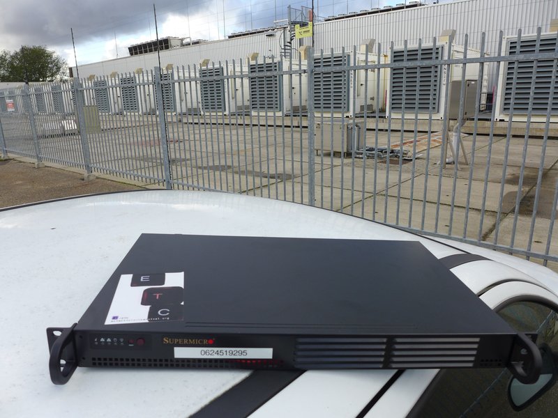 A photo of a black flat server with a sticker that has "E," "T," and "C" keyboard keys. on the roof of a car. Behind is a grey-fenced factory.