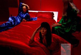 Three people with long hair wearing red, blue, and green silk dresses sit on, against, or around a red silk bed in a red room. Two of them with concerned faces gaze intently in front of them, while the other sits away.