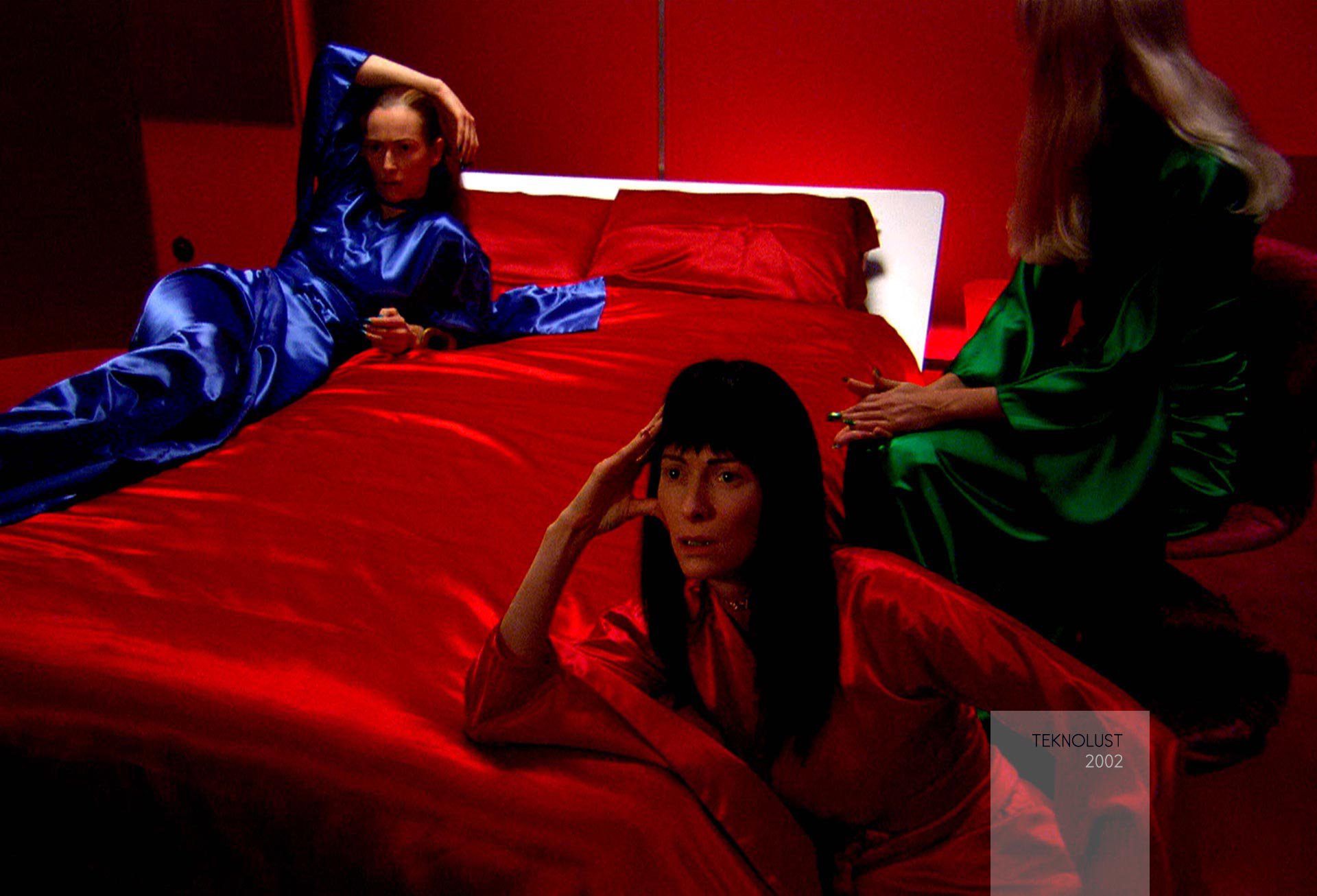 Three people with long hair wearing red, blue, and green silk dresses sit on, against, or around a red silk bed in a red room. Two of them with concerned faces gaze intently in front of them, while the other sits away.