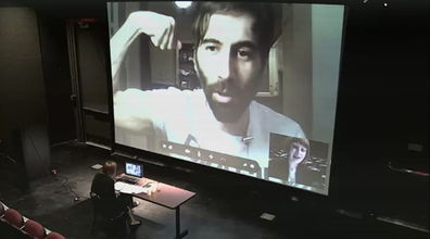 Angela Washko in a theater sits in front of a large projection of a video call of Roosh V, a white man who is flexing on camera
