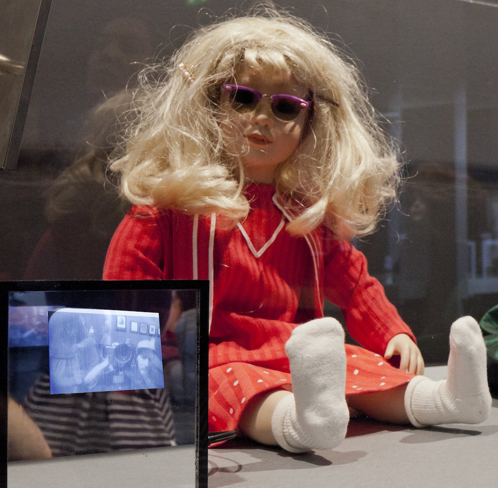 A female doll with unbrushed blonde and frizzy hair wearing a matching red outfit and white socks sits comfortably against a glass wall. Its head slightly tilts as she spies on you, expressionless, eyes covered from its sunglasses.