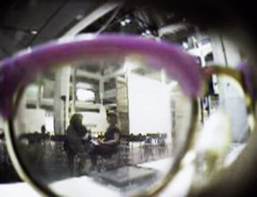 Bird's eye view of a lense looking out of a glasses frame. Two people are sitting in chairs and are deeply engaged in conversation in a vast and empty seating area with white walls, large windows, and high ceilings.