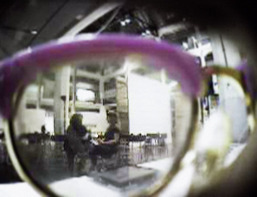 Bird's eye view of a lense looking out of a glasses frame. Two people are sitting in chairs and are deeply engaged in conversation in a vast and empty seating area with white walls, large windows, and high ceilings.