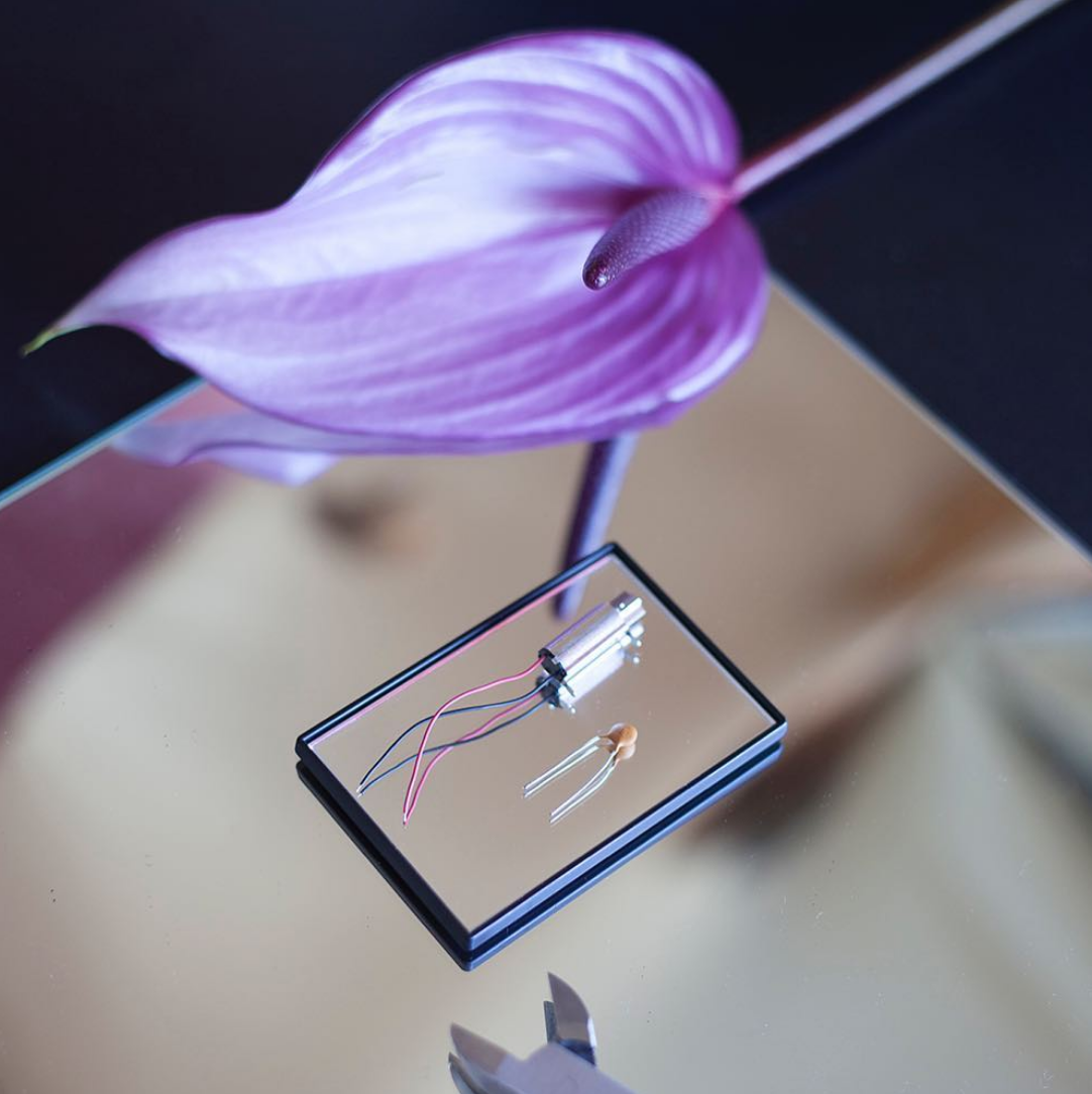 A commercial-like seductively elegant portrait of an exotic purple anthurium flower next to a metal device with red and blue wires lying next to a semiconductor device on a mirror bed stacked on a mirror plate.