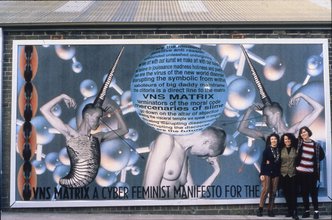 Three woman posing happily in front of the Cyberfeminist billboard that takes up most of the side of a brick building.