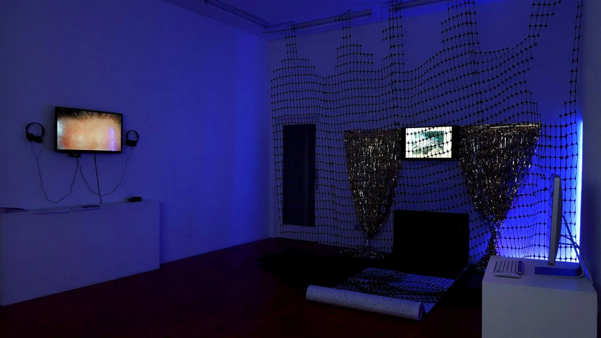 A dark gallery room is lit by blue lights. The left wall has a zoomed up forehead on a TV, the right wall has a computer on a desk, and nets in front of a TV attached to glittery curtains that fall to meet a large rolled up scroll takes up the back wall.