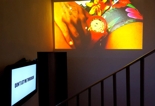 A dark room shows a stair railway leading to a white TV screen with "Don't let me through" in bold black letters. An image of a hand holding a yellow smiley face device in between thighs wearing a Hawaiian print swim suit is projected onto the right wall.